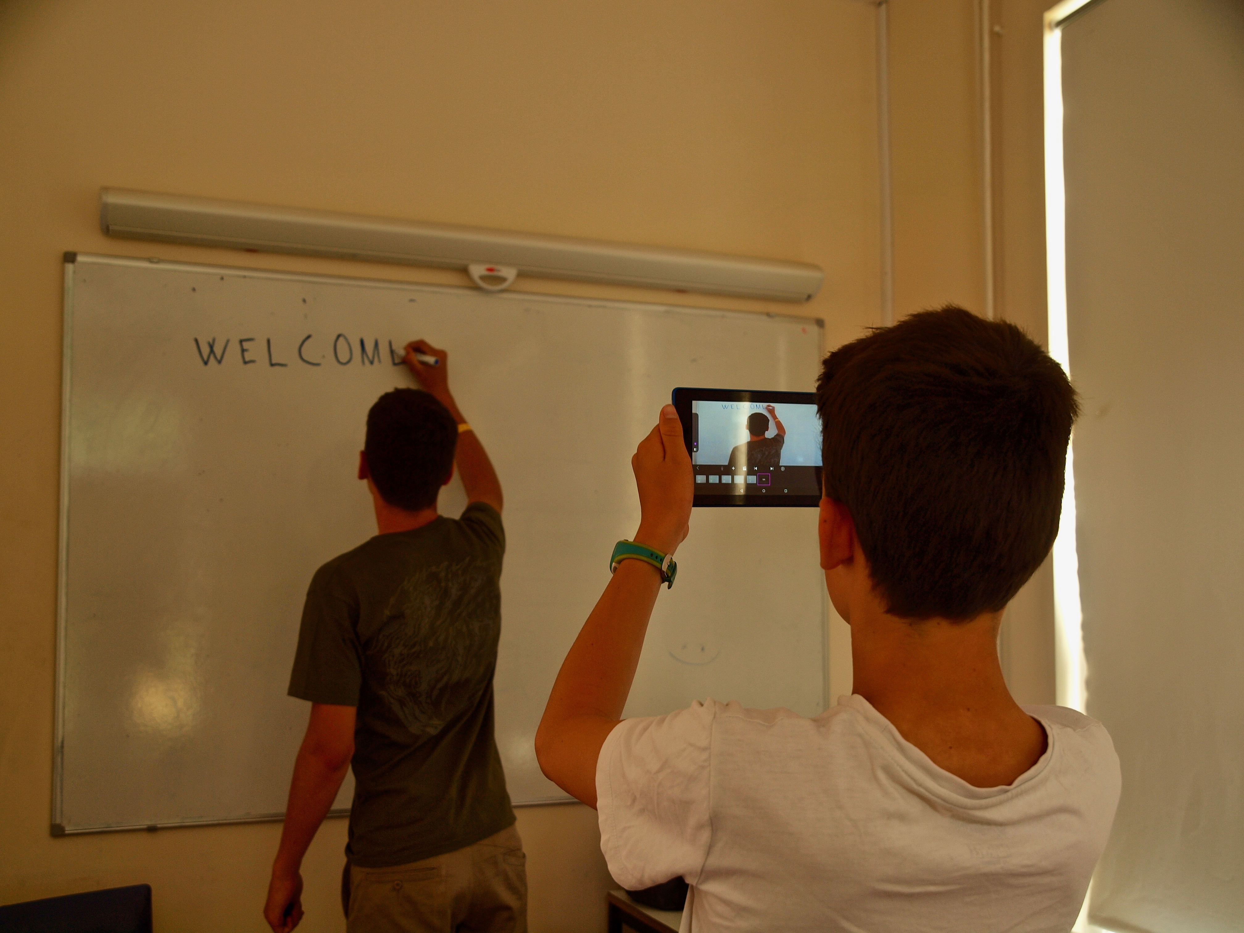 Uczeń piszący na tablicy słowo "welcome", drugi robi mu zdjęcie telefonem komórkowym