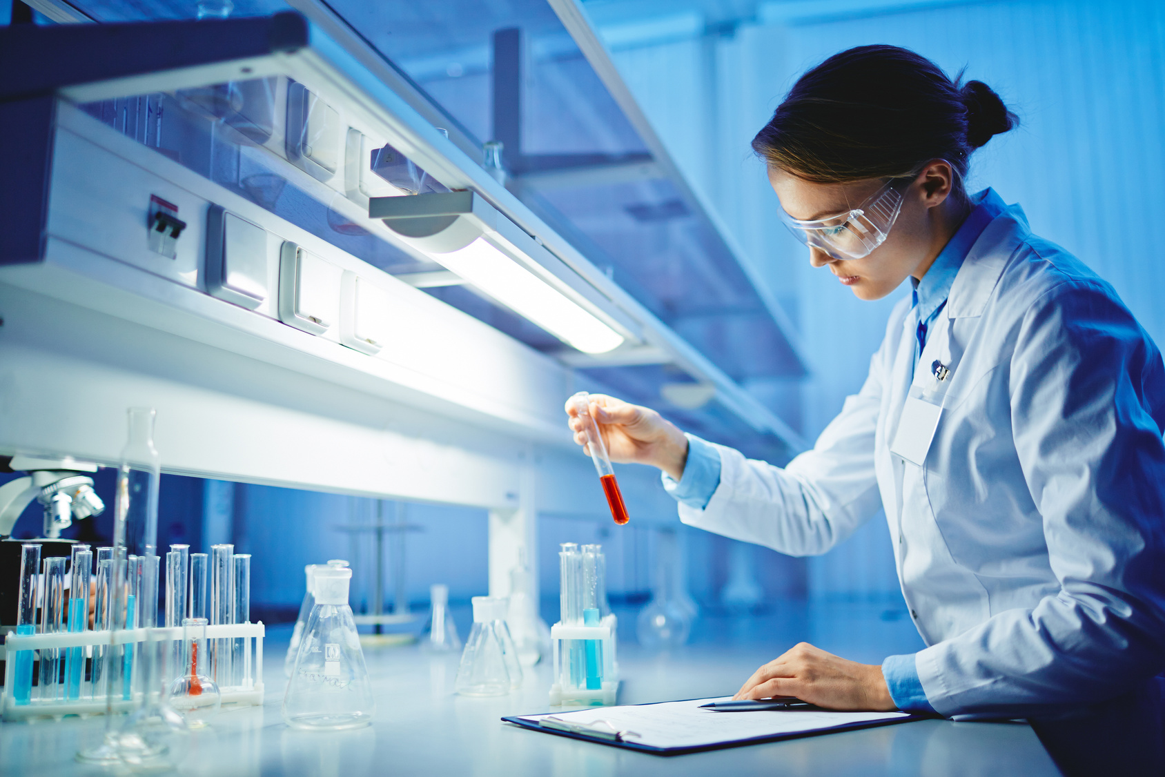 młoda kobiet w goglach ochronnych na twarzy, stoi przy blacie roboczym w laboratorium i trzyma w ręku szklaną fiolkę z różową substancją