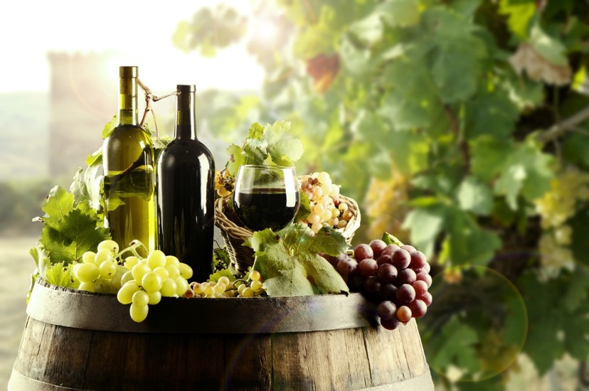 widok na butelkę wina oraz winogrona leżące na drewnianej beczce znajdującej się na tle rosnącej winorośli