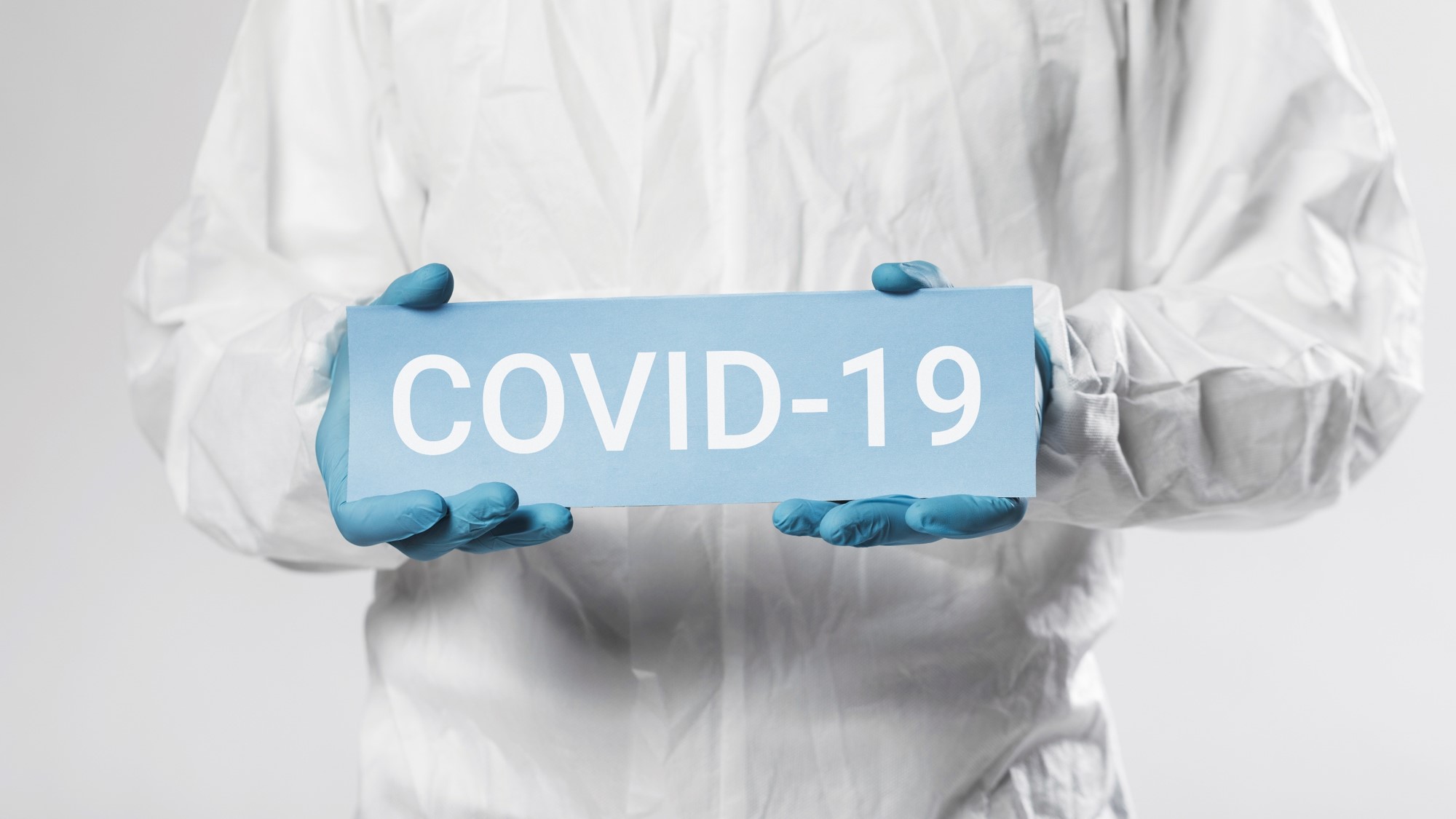 Zbliżenie na postac w kombinezonie ochronnym i plastikowych rękawiczkach, mężczyzna trzyma w rękach tabliczkę z napisem: Covid-19