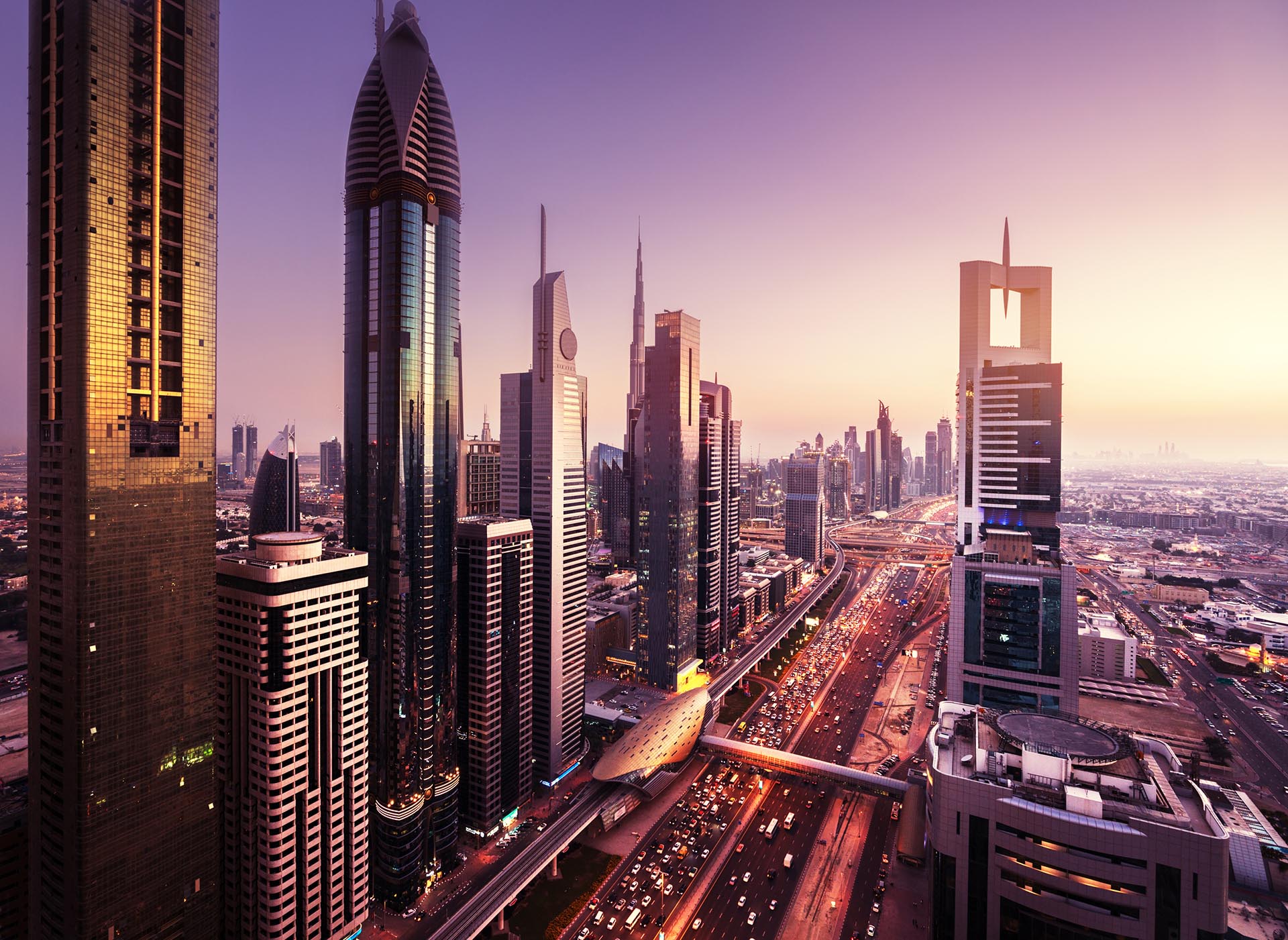 widok z lotu ptaka podczas zachodu słońca na ulicę, przy której stoją liczne smukłe wieżowce o szklanych elewacjach. To miasto to Dubaj