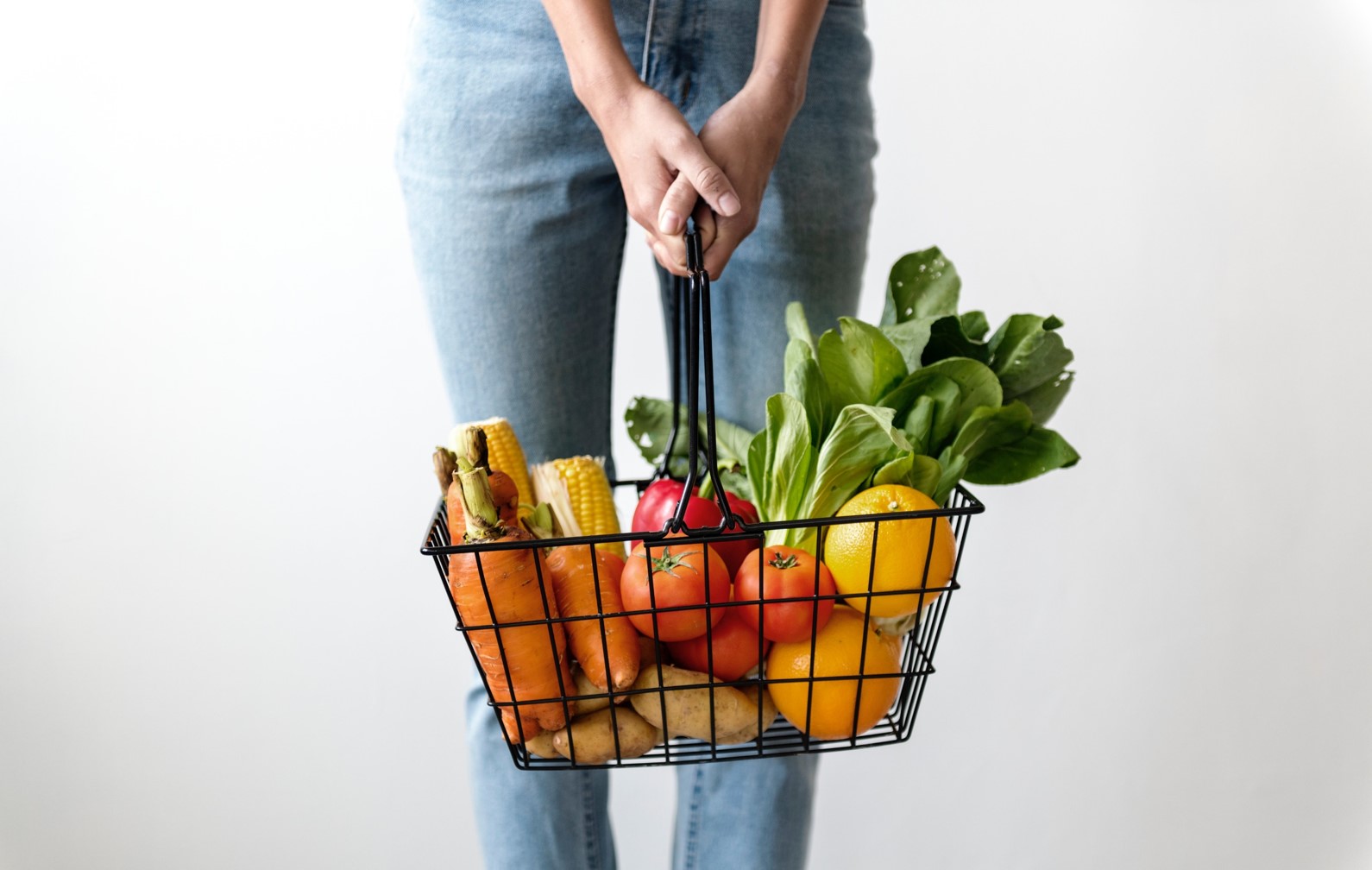 Widok od pasa w dół na kobietę w jeansach trzymającą oburącz ażurowy koszyk zakupowy wypełniony owocami i warzywami