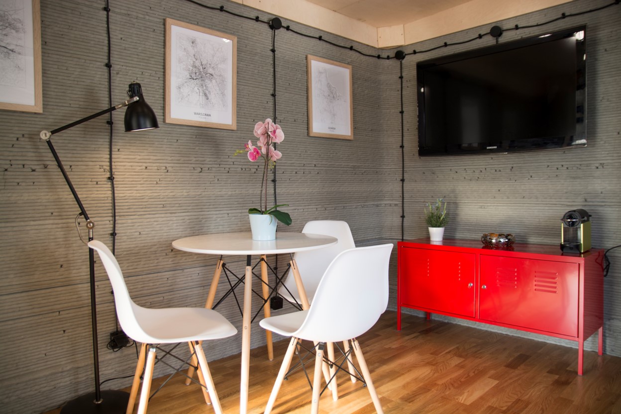 Widok na wnętrze niewielkiego pomieszczenia, w którym znajdują się stół, krzesła, komoda w czerwonym kolorze, stojąca na podłodze lampa oraz wiszące na ścianach obrazki i telewizor. Podłoga jest wykończona drewnem. Ściany są w naturalnym kolorze betonu.