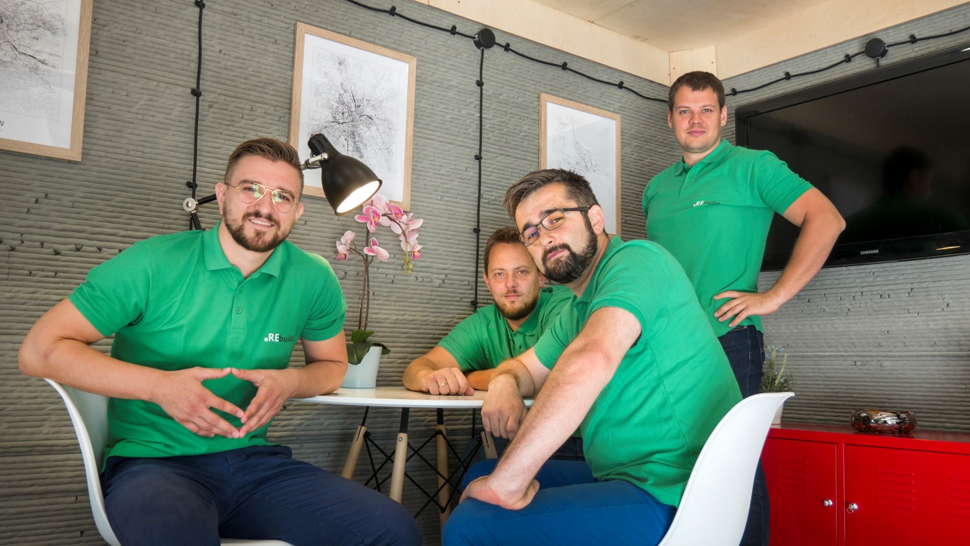 Widok na czterech mężczyzn w średnim wieku znajdujących się w niewielkim pomieszczeniu. Trzech z nich siedzi, jeden zaś stoi. Wszyscy mają zielone koszuli i wpatrują się w obiektyw. 